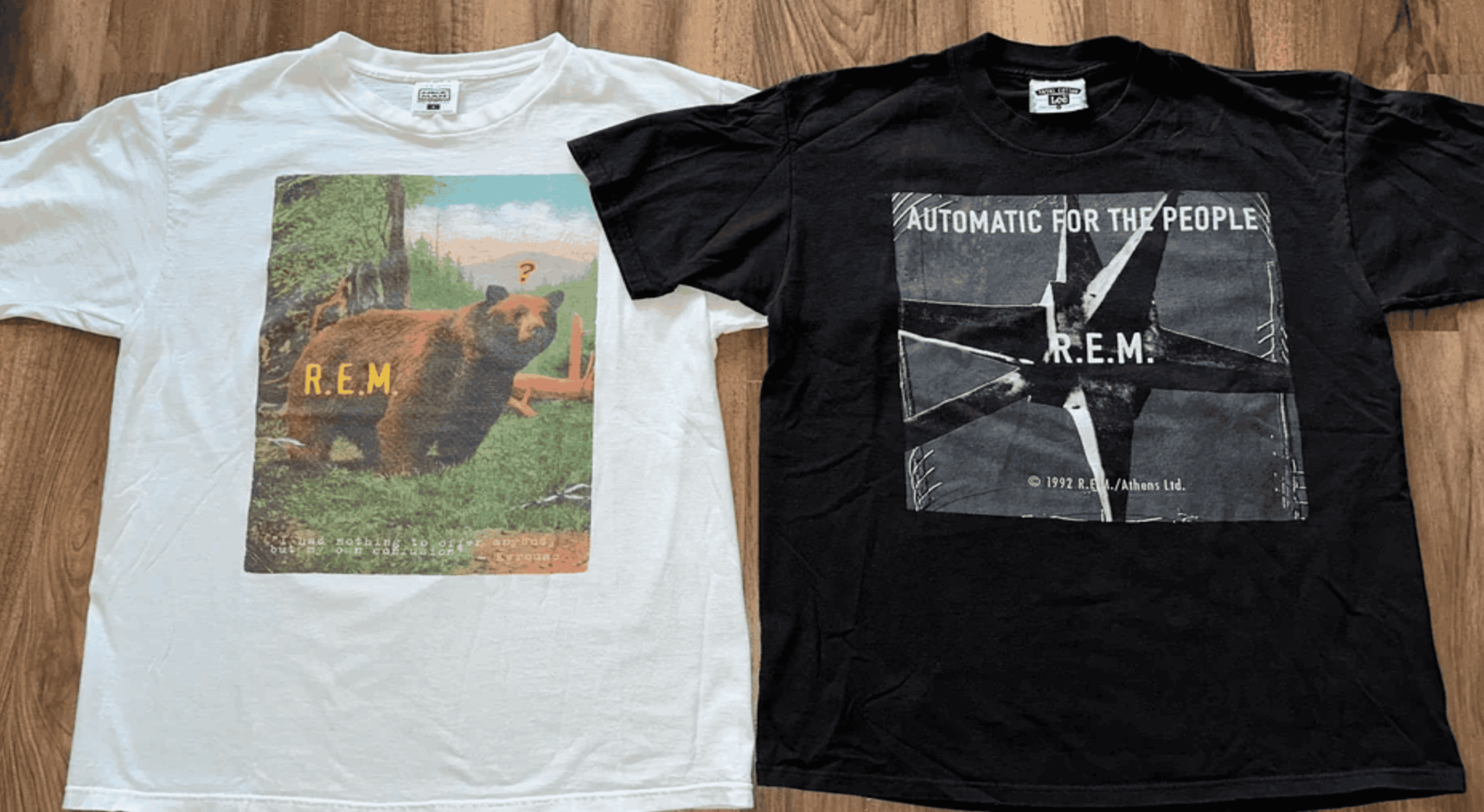 Two R.E.M. band t-shirts on a wooden floor; the white shirt shows a bear and the black shirt features the “Automatic for the People” album cover.
