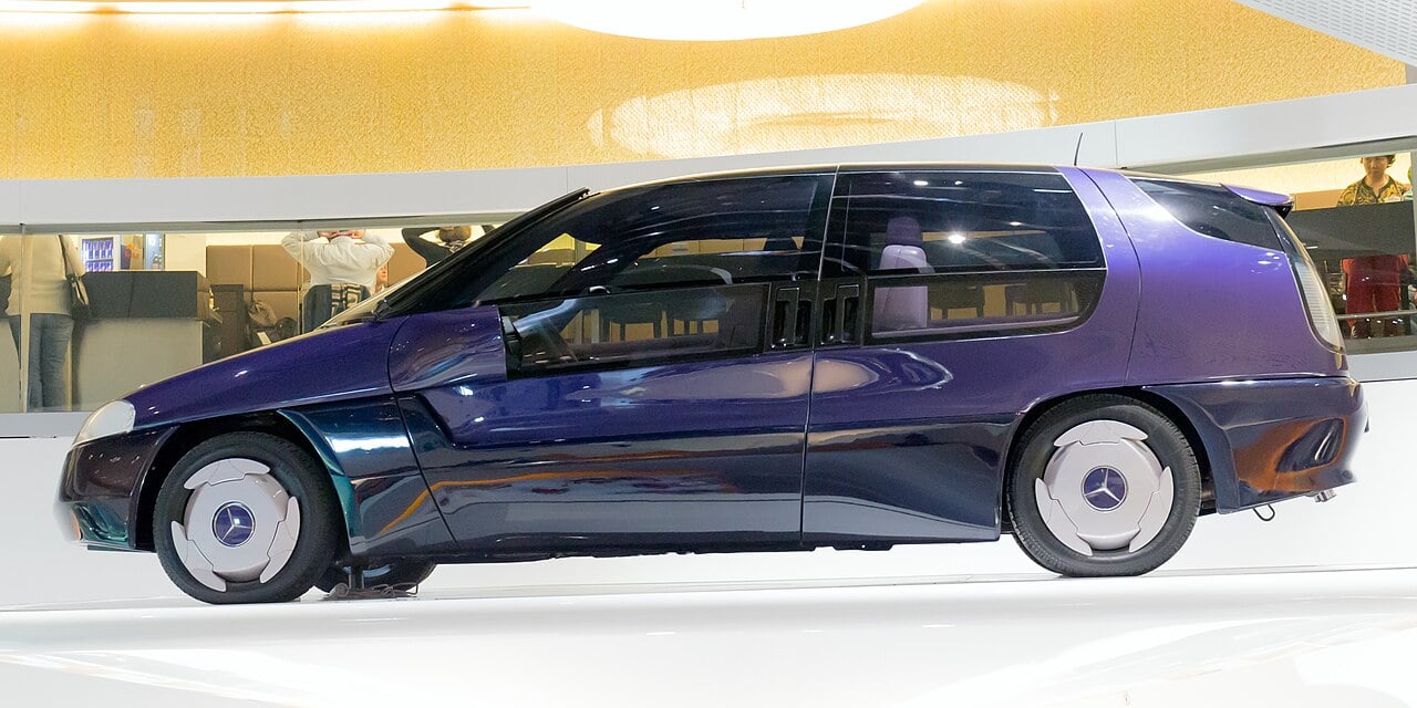 A futuristic, purple Mercedes-Benz concept car with a sleek, aerodynamic design is displayed indoors under bright lighting. The vehicle features large windows and unique wheels.