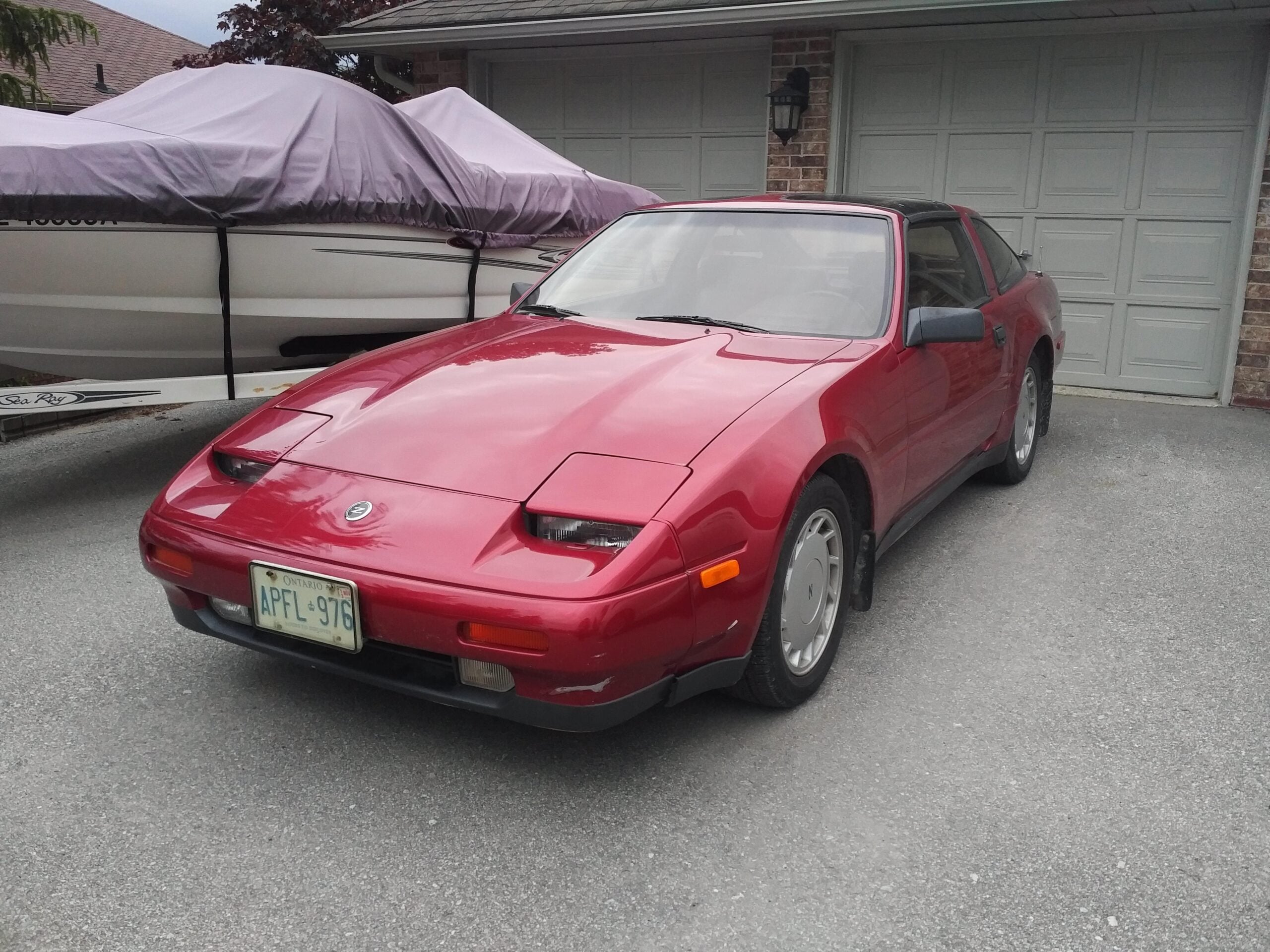 A red sports car with pop-up headlights is parked in front of a gray garage door next to a covered boat on a trailer.