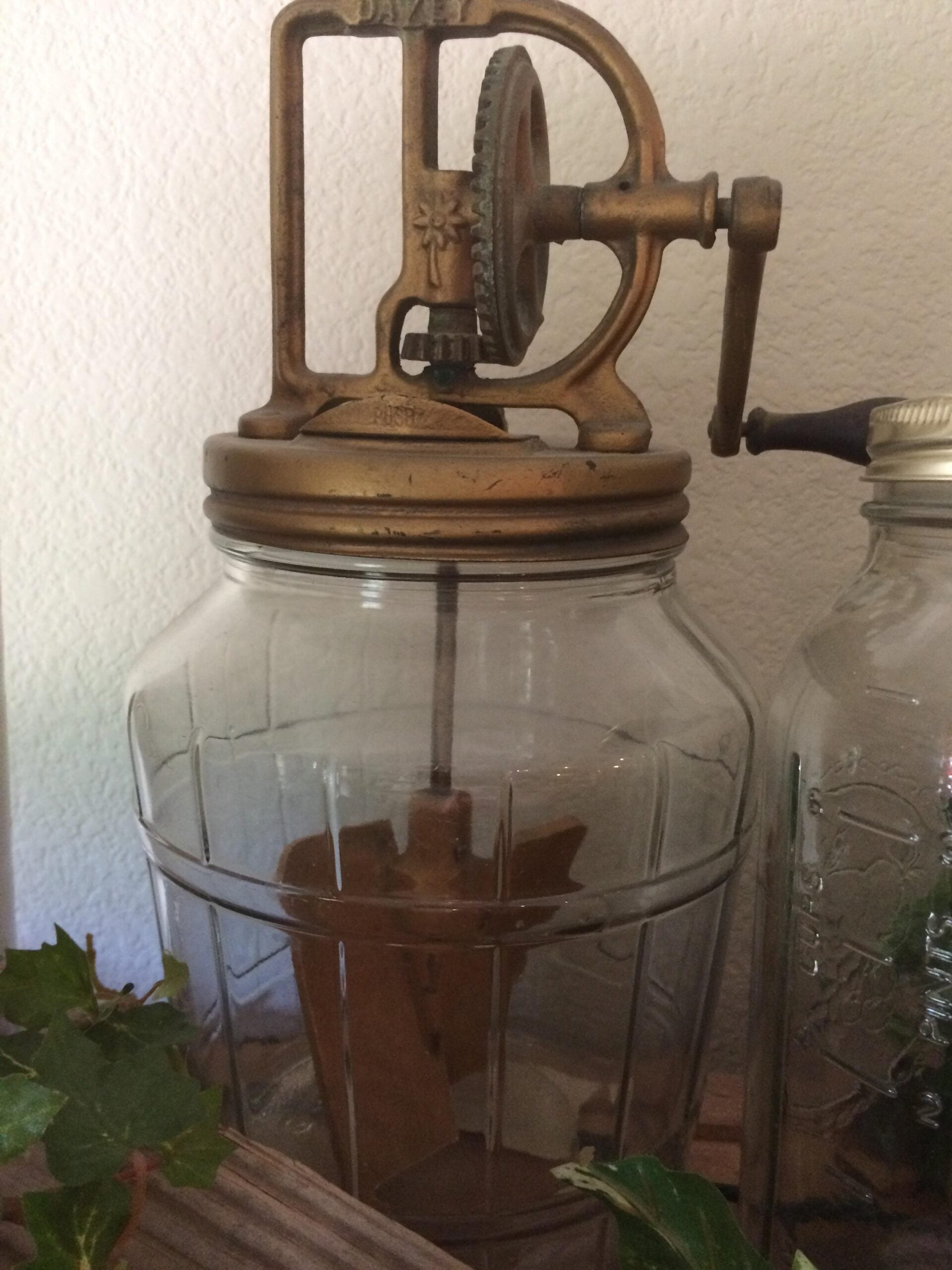 A vintage glass butter churn with a metal lid and hand-crank mechanism sits on a wooden surface next to a mason jar. Some green leaves are visible in the foreground.