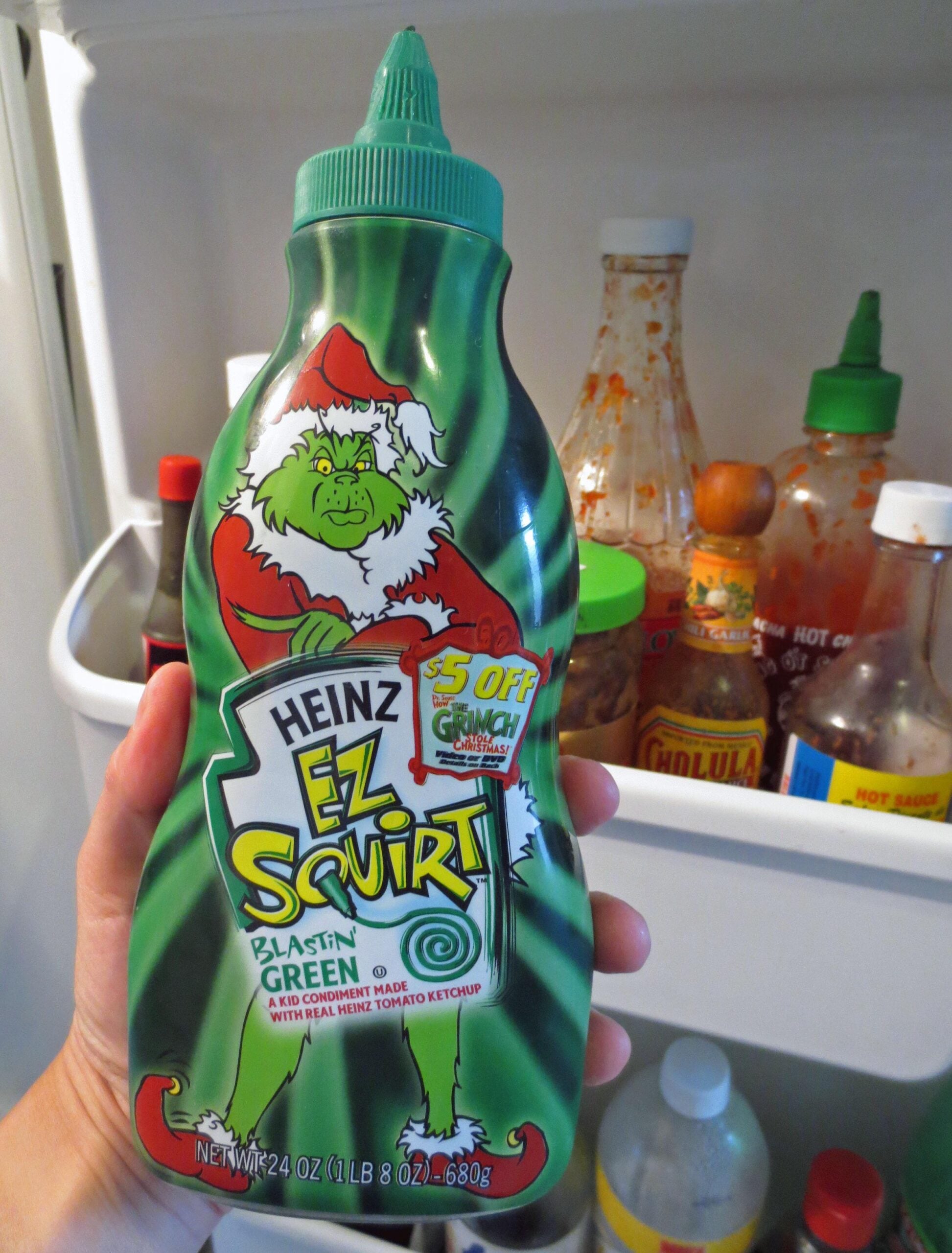 A hand holds a green Heinz EZ Squirt ketchup bottle featuring the Grinch, with assorted sauces visible in the refrigerator door behind it.