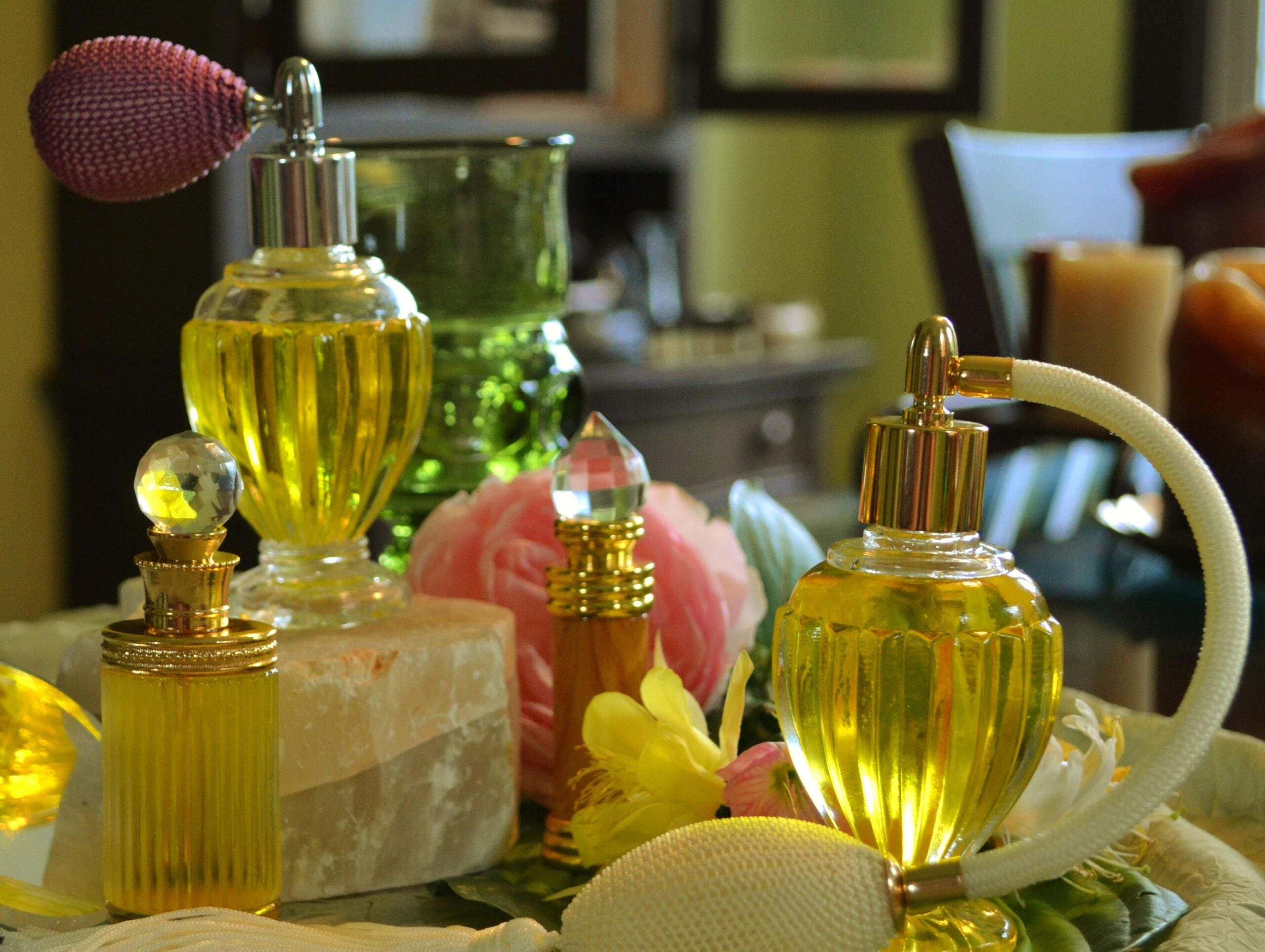 Vintage glass perfume bottles with yellow liquid are arranged on a table with a pink rose, a yellow flower, and candles in the background. The scene has a soft, elegant, and vintage feel.
