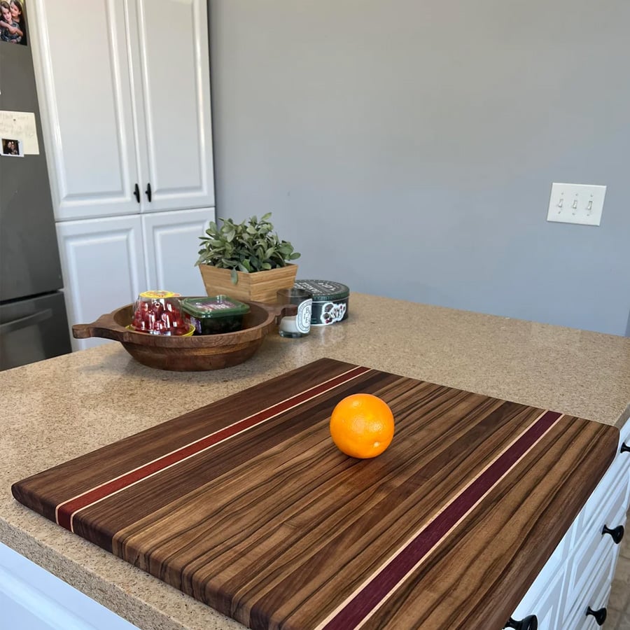 A single orange sits on a striped wooden cutting board on a kitchen counter, with bowls and a potted plant in the background.