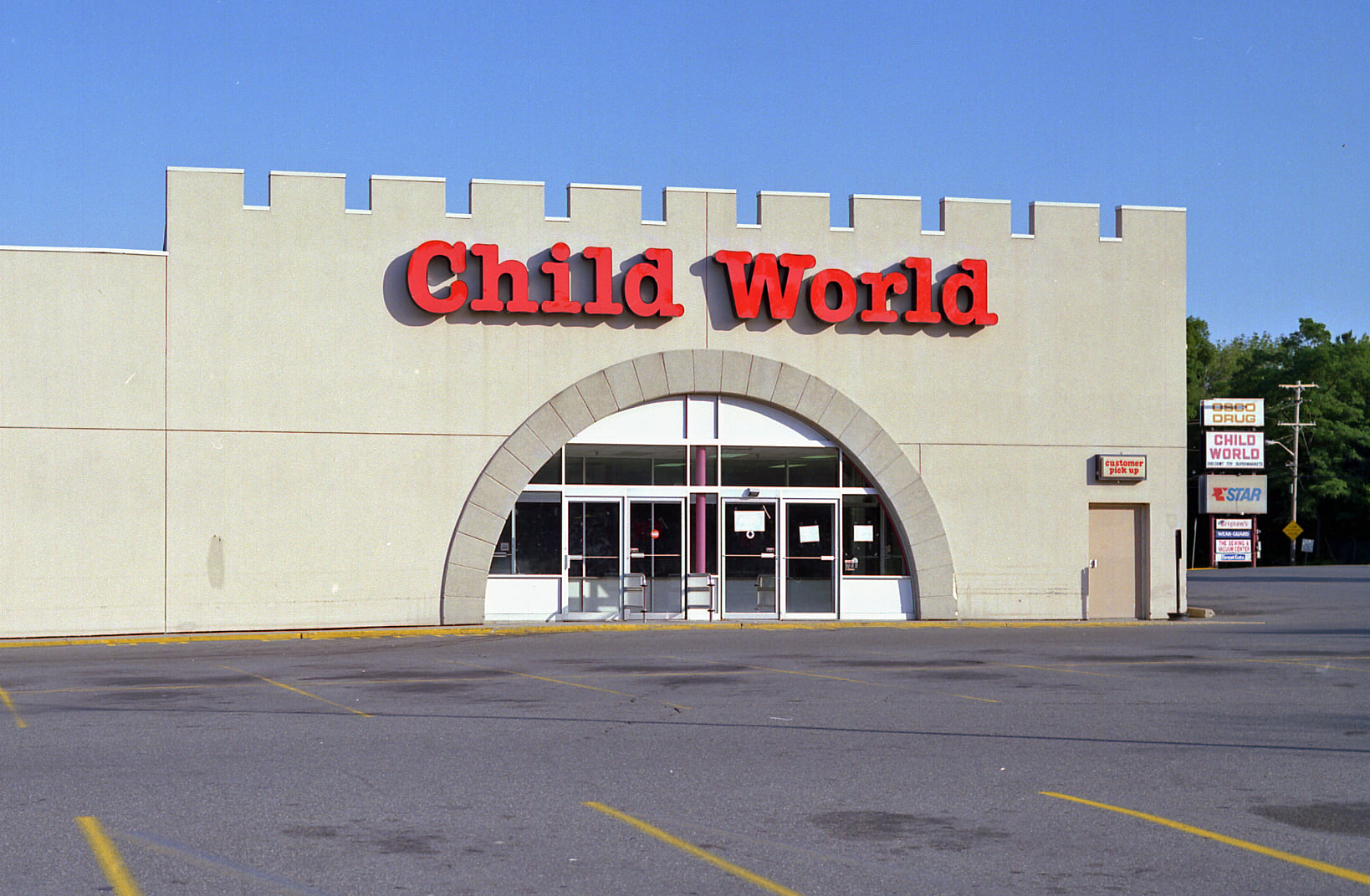 A storefront with a castle-like facade displays the red sign “Child World.” The building has an arched entrance and the parking lot is mostly empty. A sign with store hours stands near the entrance.