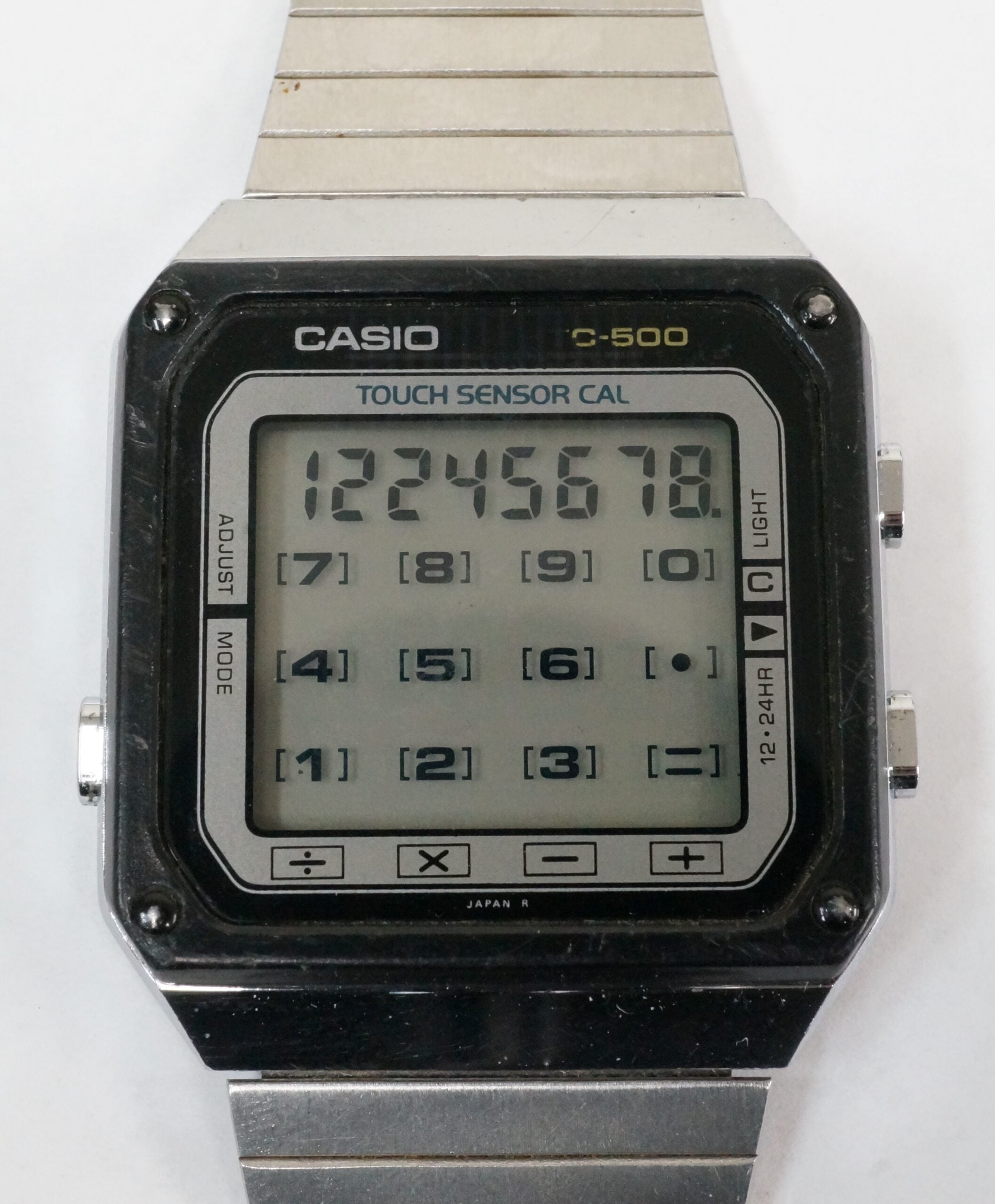 A close-up of a Casio C-500 digital watch with a touch sensor calculator, displaying numbers 1 to 8 on the screen above a keypad grid. The watch has a metal band and a rectangular face.