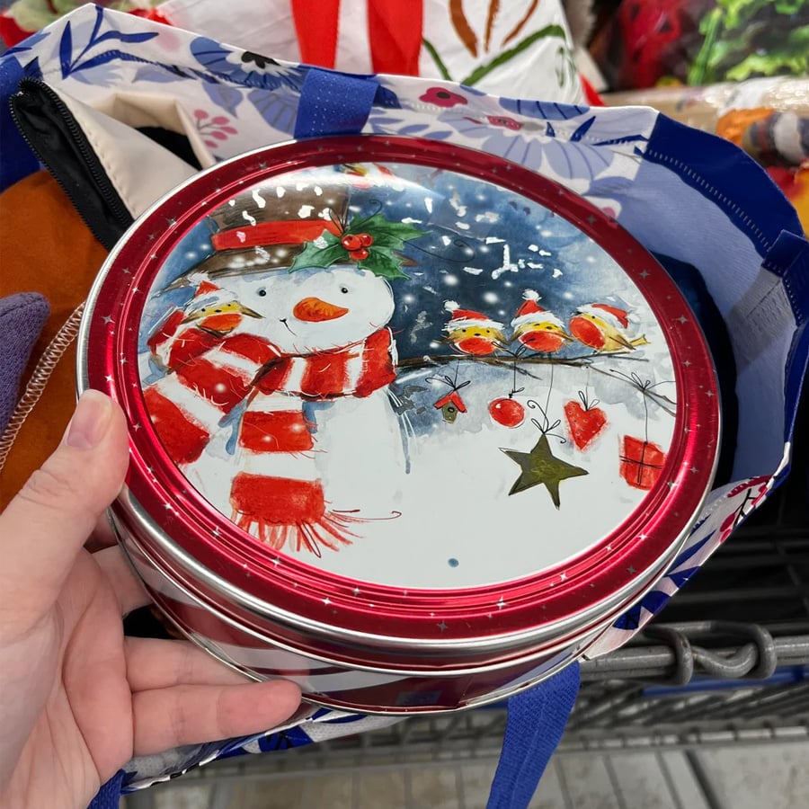 A hand holds a metal tin with a festive snowman, red scarf, hat, and three birds on a snowy background. Musical notes and ornaments decorate the lid. The tin is inside a shopping bag.