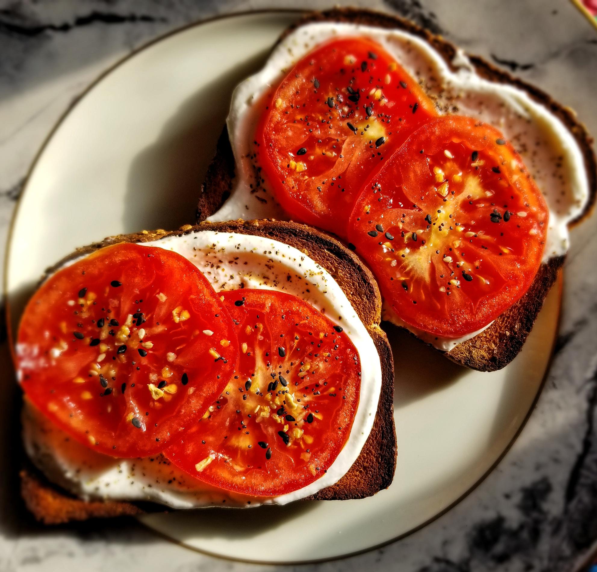 Two slices of toast topped with cream cheese, fresh tomato slices, and a sprinkle of seasoning on a white plate with a gold rim, set on a marble surface.
