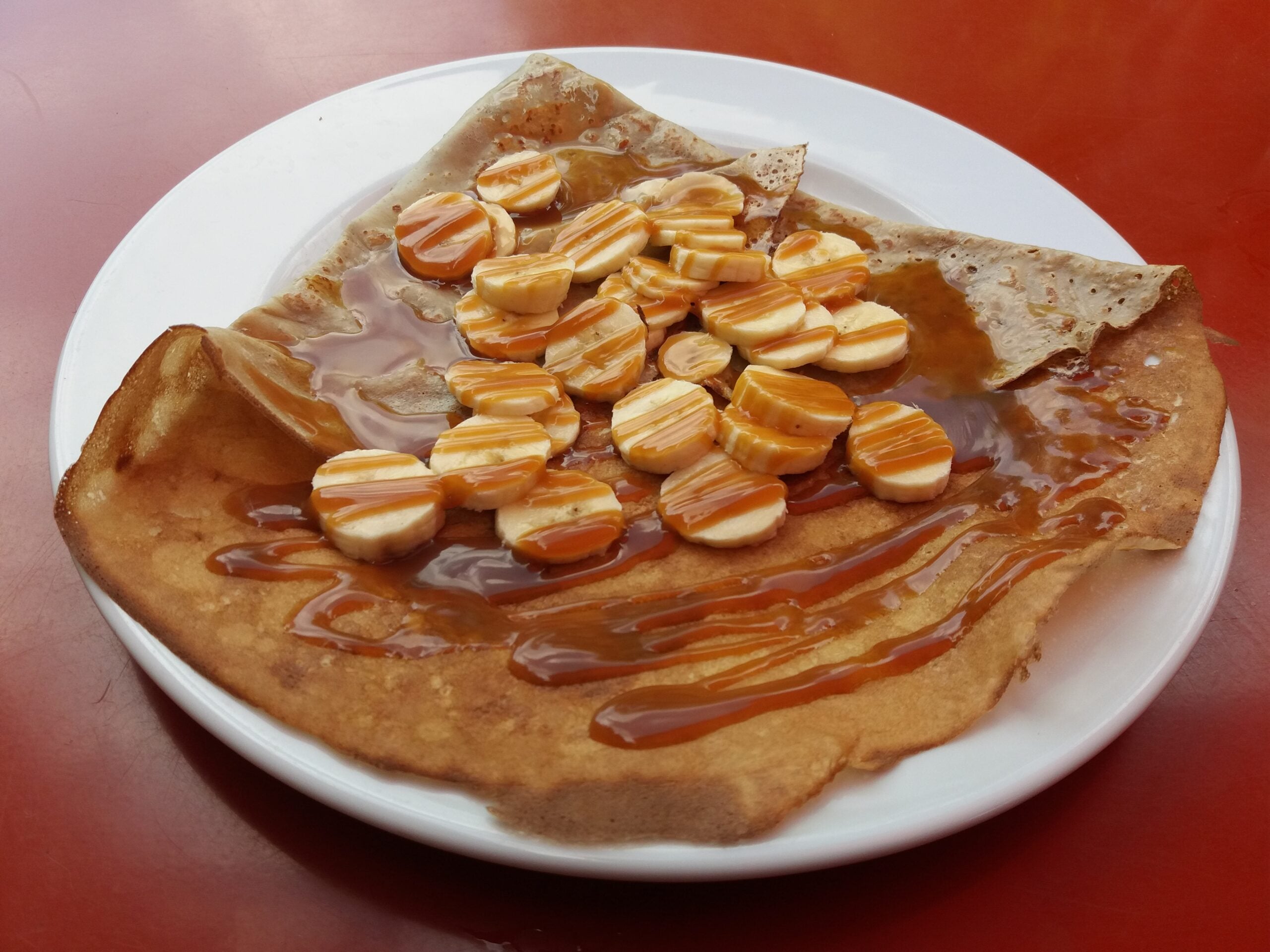 A crepe on a white plate topped with sliced bananas and drizzled with caramel sauce, set on a red table.