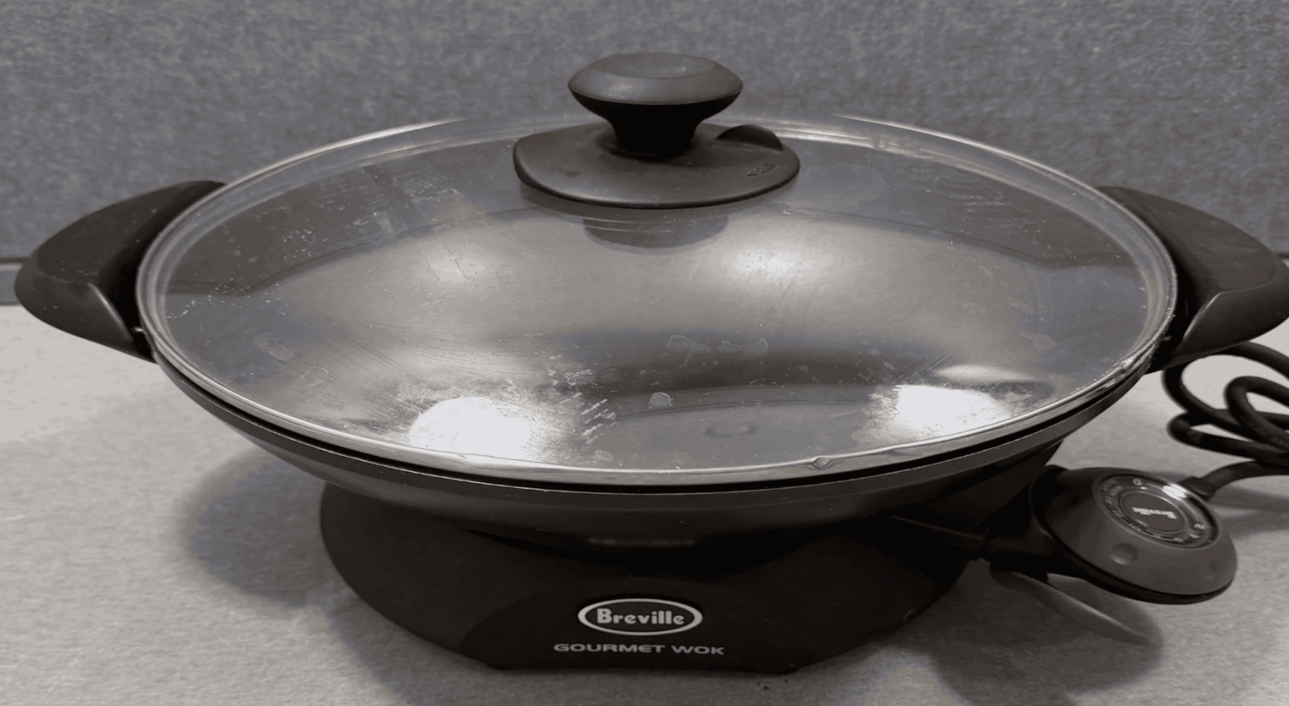 A black Breville electric gourmet wok with a glass lid and side handles sits on a gray surface. The wok is plugged in, and the temperature control is visible on the right side.