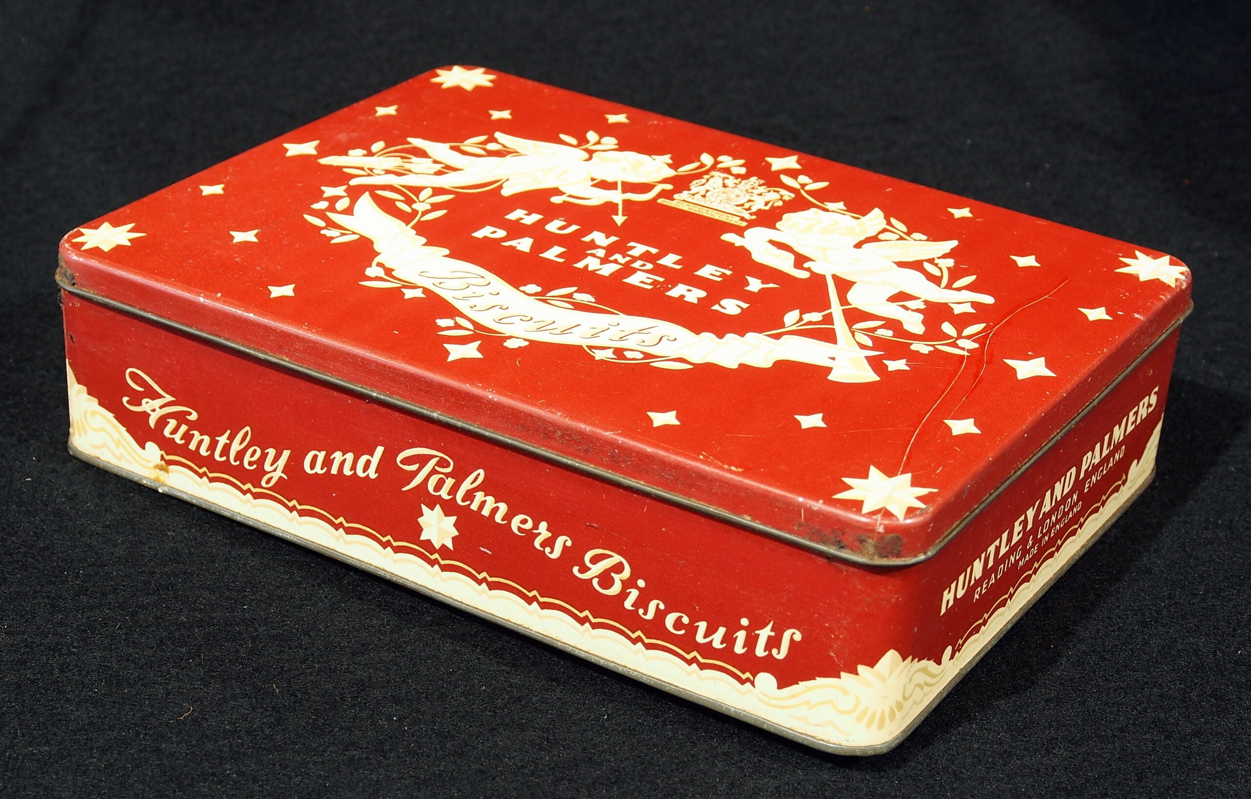 A red Huntley & Palmers Biscuits tin box with white decorative designs and two cherubs holding a wreath on the lid, set against a black background.