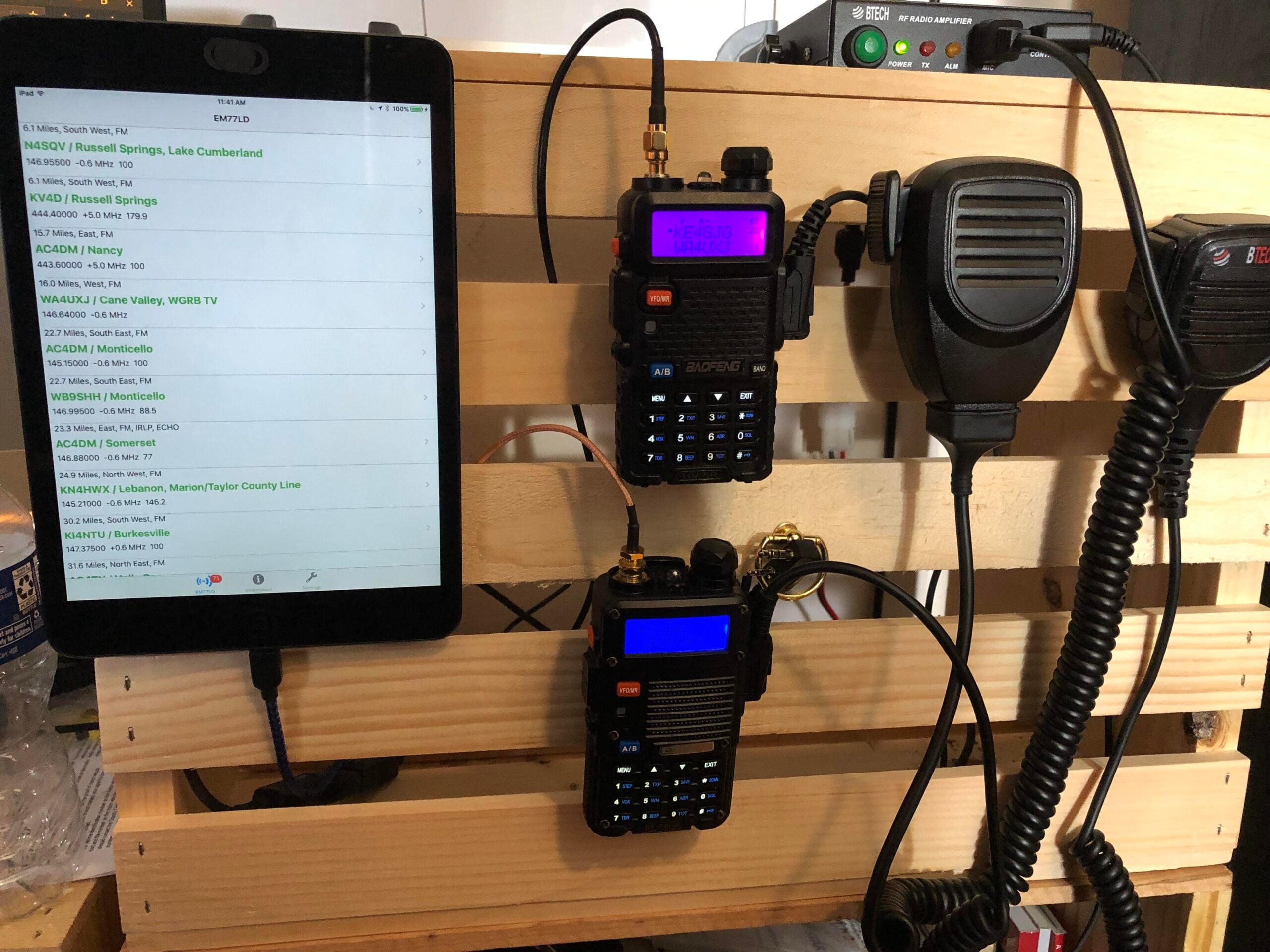 A wooden crate holds two-way radios, microphones, cables, and a tablet displaying a list of radio frequencies or call signs. Devices are neatly arranged, suggesting a radio communication setup.