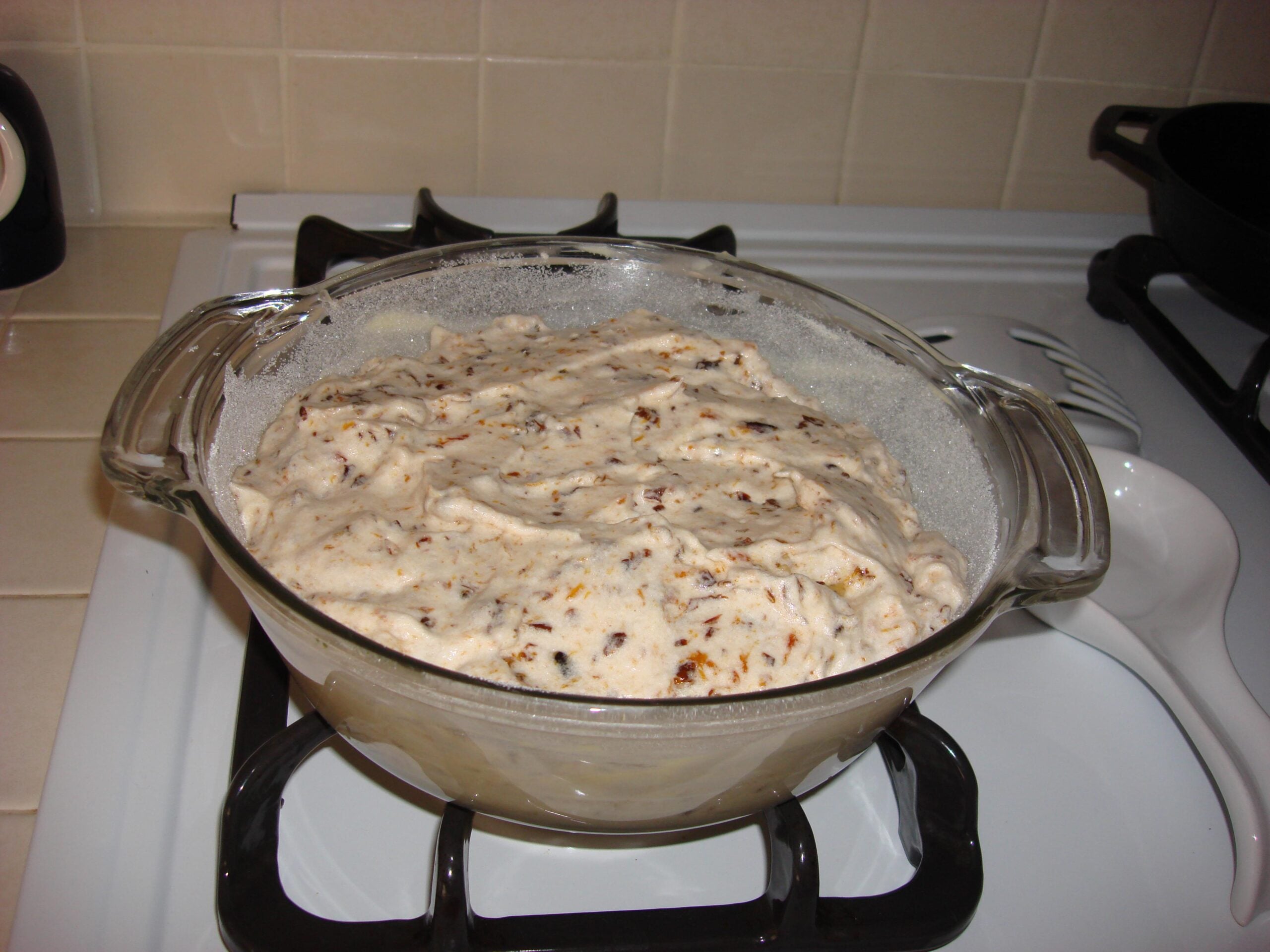A glass bowl filled with ice cream mixture, containing chocolate chunks, sits on a stovetop in a kitchen with beige tiled walls.