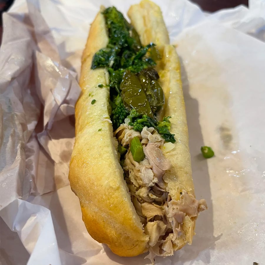 A sandwich on a hoagie roll filled with shredded meat, sautéed greens, and sliced green peppers, served on crinkled white paper.