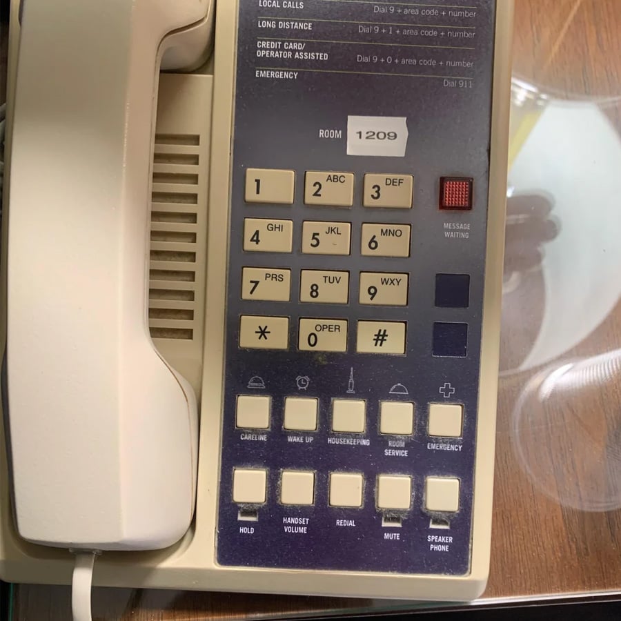 A beige corded hotel phone with a numeric keypad, labeled buttons, and a display showing room “1209” sits on a wooden surface next to a partially visible plate.