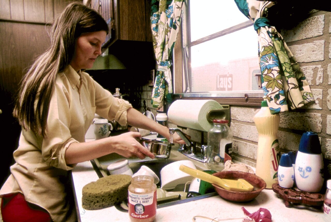 A woman washes dishes at a kitchen sink beside a window with floral curtains. Various kitchen items, including a sponge, dish soap, and a jar of baby food, are on the countertop.