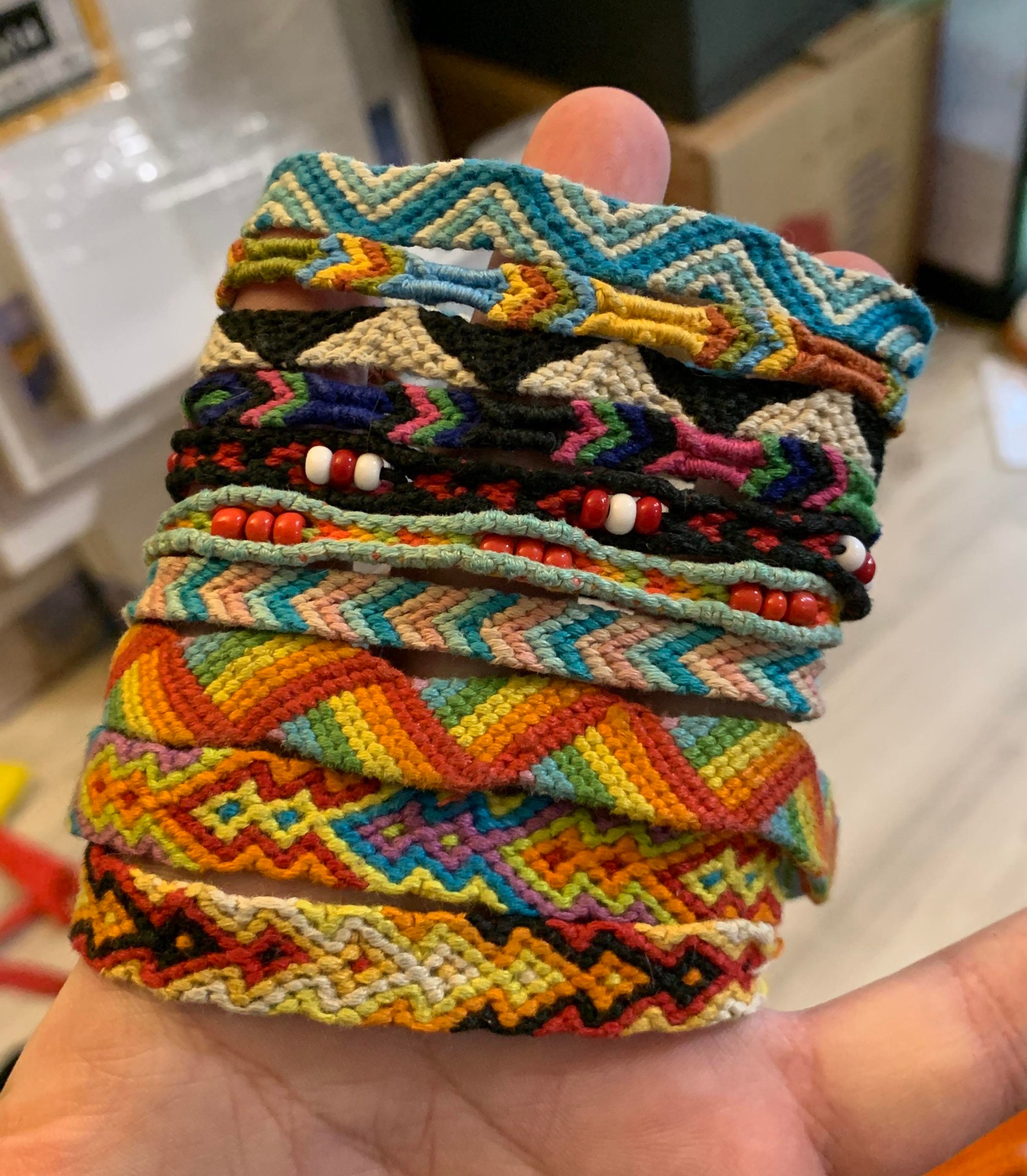 A hand holds several woven friendship bracelets with colorful, intricate patterns and bead accents. The background is slightly blurred, showing boxes and a wooden floor.
