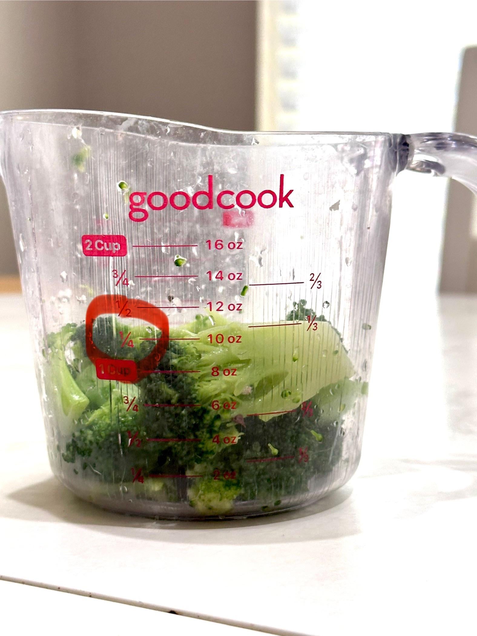 A clear measuring cup labeled “goodcook” with chopped broccoli inside, measuring about 1 cup. The cup has red measurement markings for ounces, cups, and fractions.