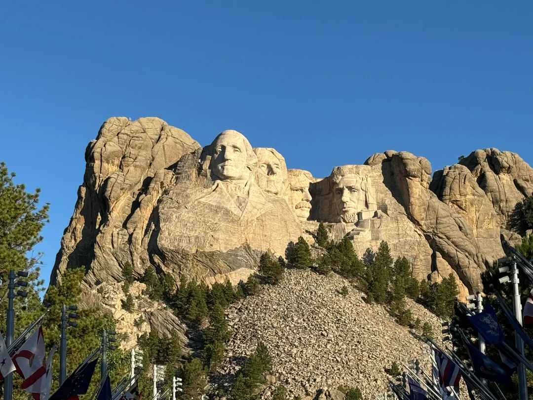 Mount Rushmore National Memorial with four presidents’ faces carved into a granite mountain, surrounded by trees, clear blue sky, and flags in the foreground.