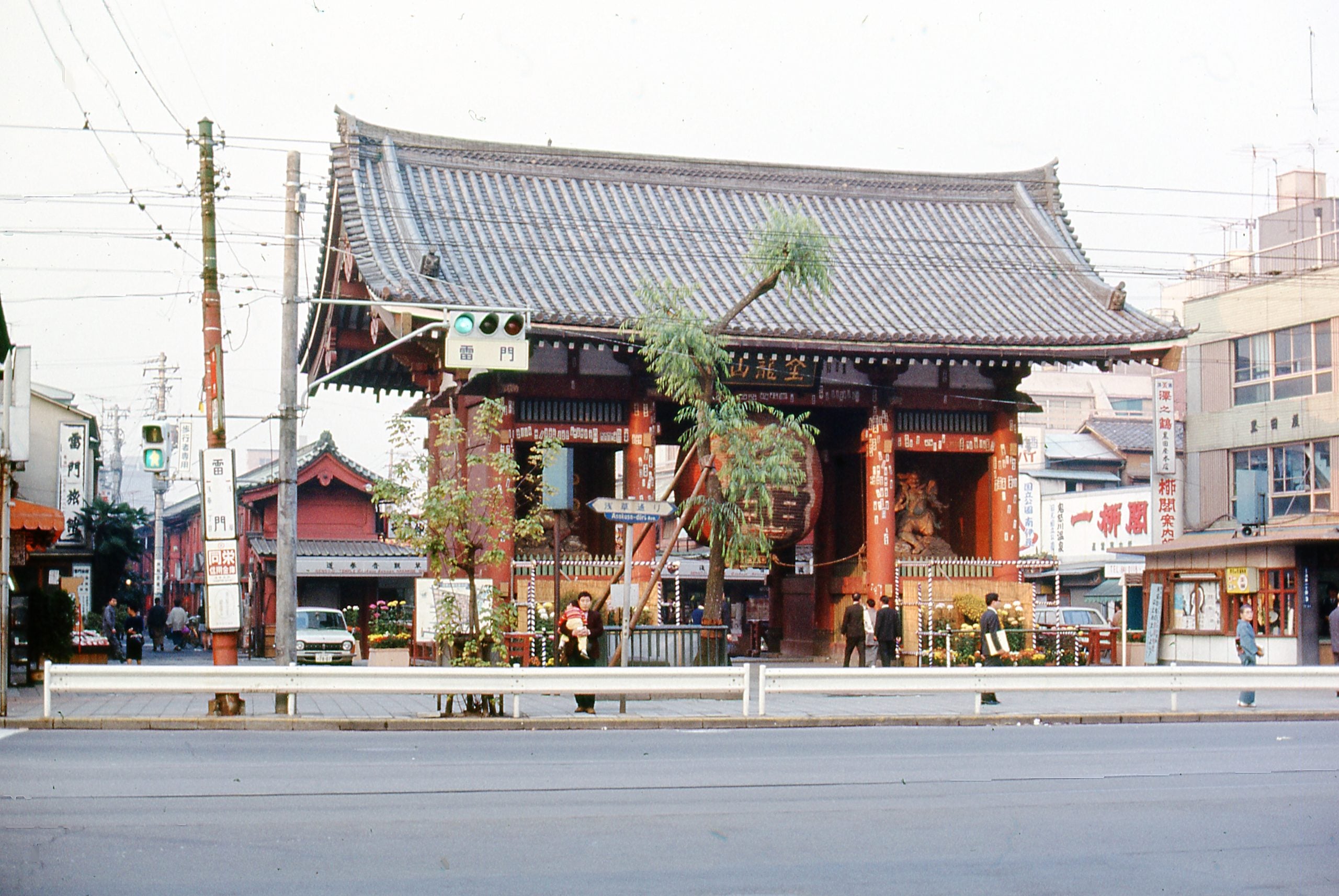 A traditional Japanese temple gate stands by a busy street with people walking nearby, flanked by shops and buildings, under a cloudy sky.