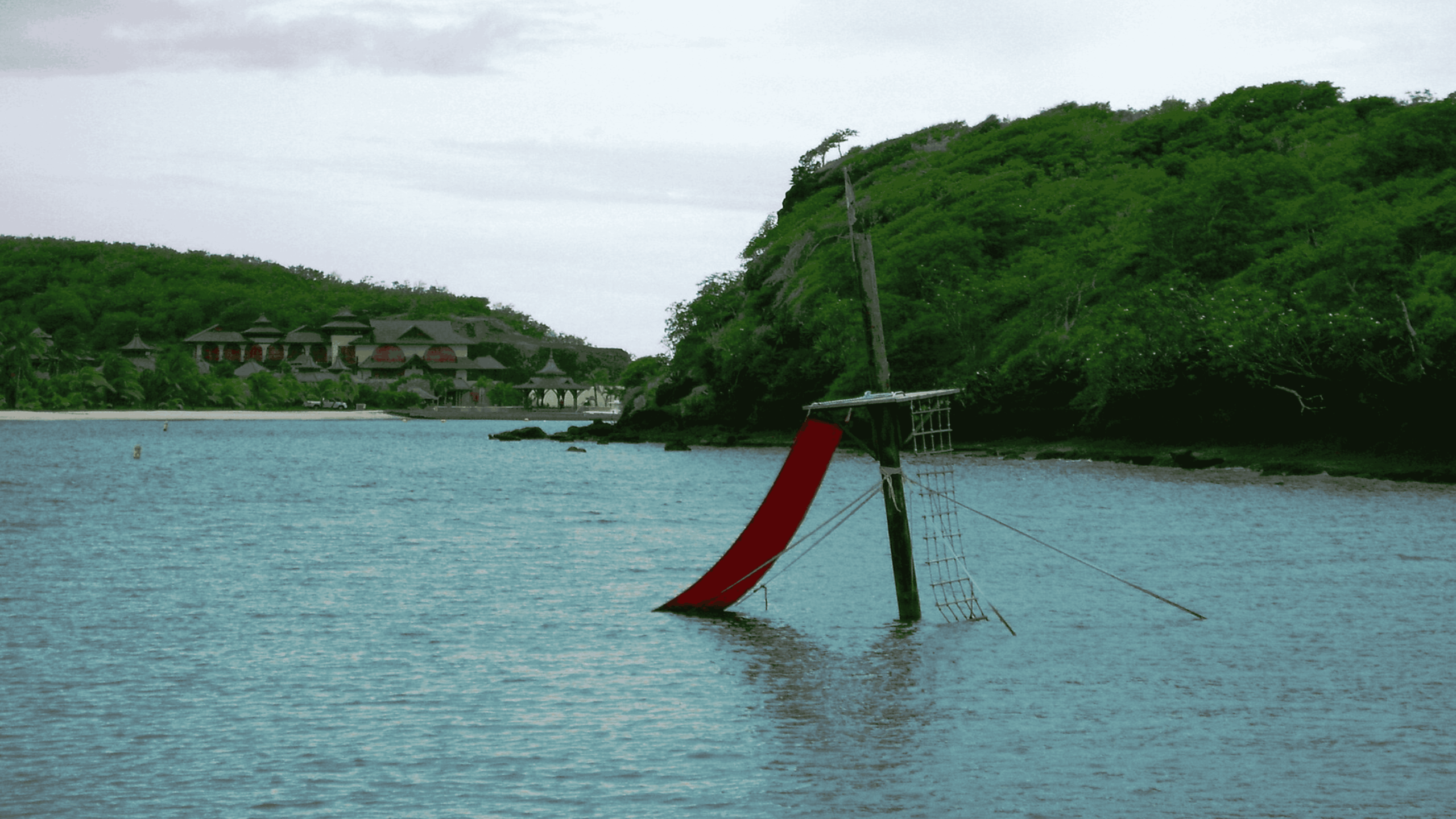 A partially sunken ship mast with a red sail rises from calm water near a lush, green shoreline and distant buildings under a cloudy sky.