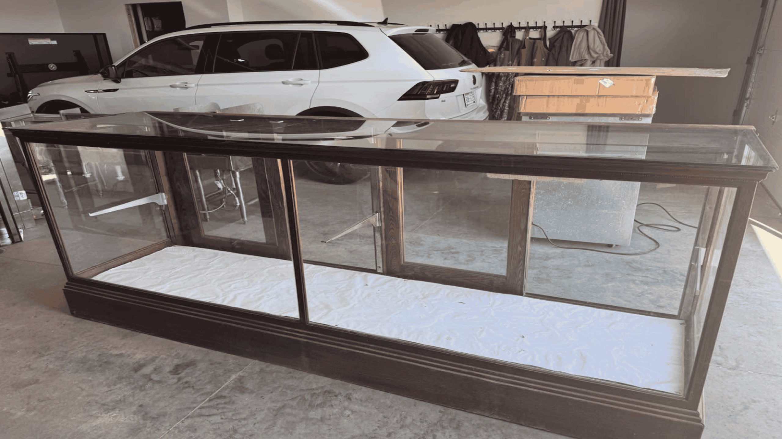 A large glass display case with dark wood frame stands in a garage. A white SUV and some boxes are in the background, along with a coat rack and other miscellaneous items.
