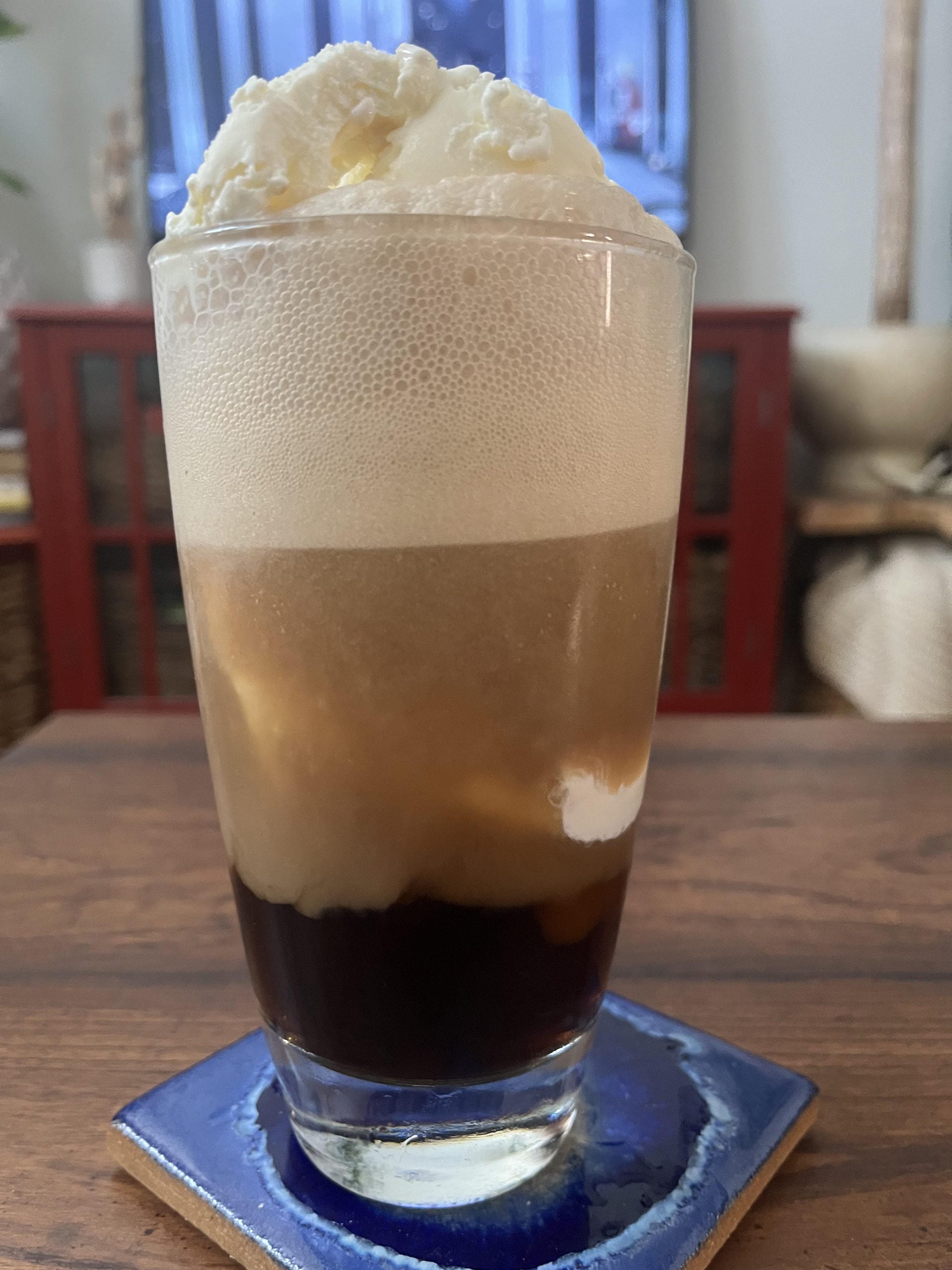A glass of root beer float with vanilla ice cream sits on a blue coaster atop a wooden table, with a blurred background of furniture and decor.