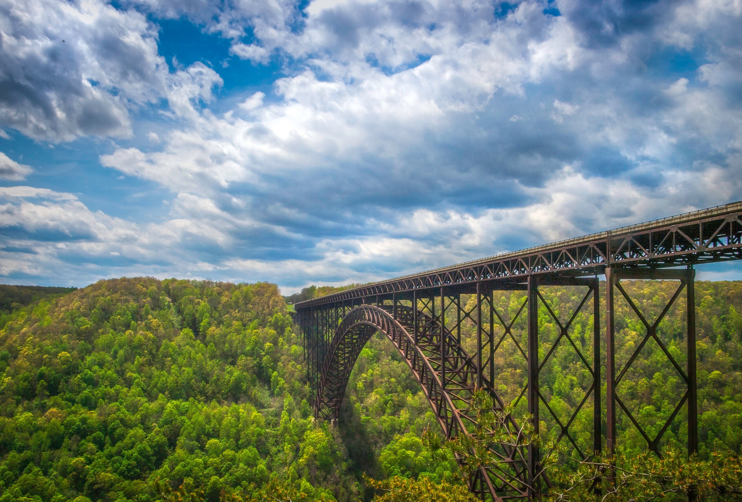 Large steel arch bridge spans a lush green forest under a partly cloudy blue sky, with rolling hills covered in trees visible below and beyond the bridge.