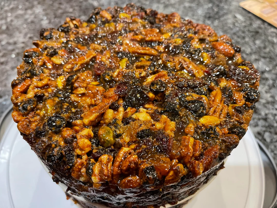 A close-up of a round fruitcake topped with glistening mixed nuts and dried fruit, resting on a white plate with a dark granite countertop in the background.