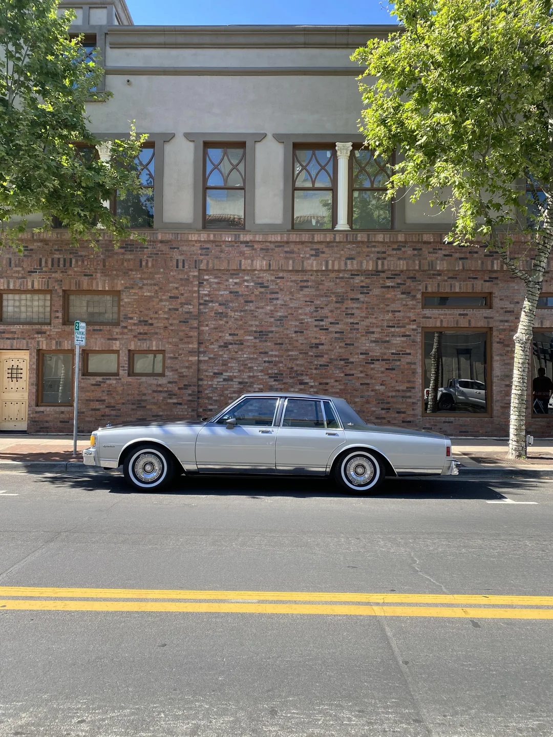 A silver classic car is parked on a street in front of a building with brick and tan walls, tall windows, and trees providing partial shade.