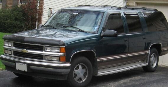 A dark green Chevrolet Suburban SUV is parked in a driveway beside a house, with some grass visible on the left and part of a garage on the right. The vehicle has four doors and roof rails.