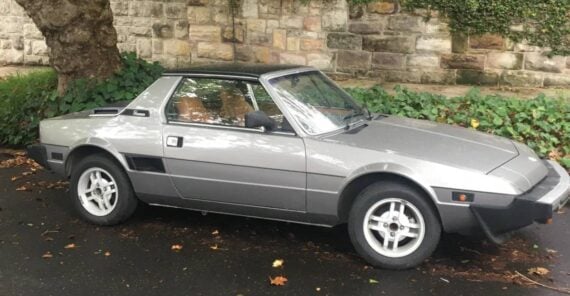 A silver classic sports car with a black roof is parked on a wet street near a stone wall, green ivy, and large trees.