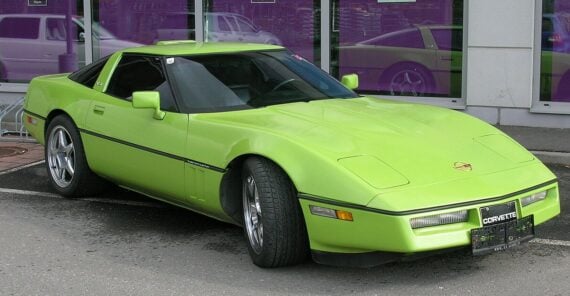 A bright green Chevrolet Corvette sports car is parked in front of a building with large purple-tinted windows reflecting another Corvette.