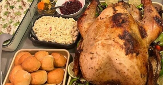 A roasted turkey on a platter with limes, surrounded by dinner rolls, macaroni salad, cranberry sauce, and a casserole topped with herbs.