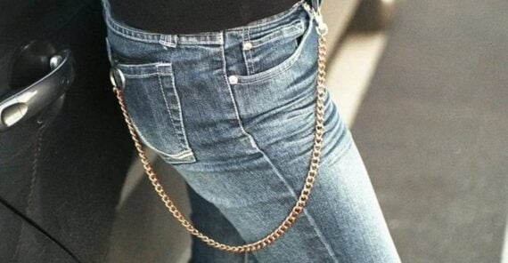Person wearing blue jeans with a gold chain wallet attached to a belt loop, leaning against a black car. Only the lower torso and upper legs are visible.