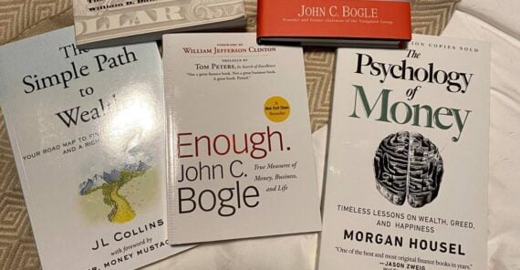 Five personal finance books are arranged on a bed, including “The Millionaire Next Door,” “The Little Book of Common Sense Investing,” “The Simple Path to Wealth,” “Enough,” and “The Psychology of Money.”