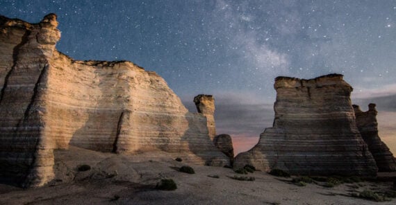 Tall, rugged rock formations rise from a sandy landscape under a star-filled night sky, with the Milky Way faintly visible above the cliffs.