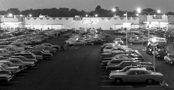Black and white photo of a crowded 1960s parking lot at night, filled with classic cars, in front of a large store with a sign reading “Two Guys.” Streetlights illuminate the area.