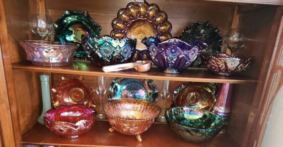 Five colorful glass vases with angular, geometric patterns in shades of green, blue, and purple are displayed on a light wooden surface against a white background.
