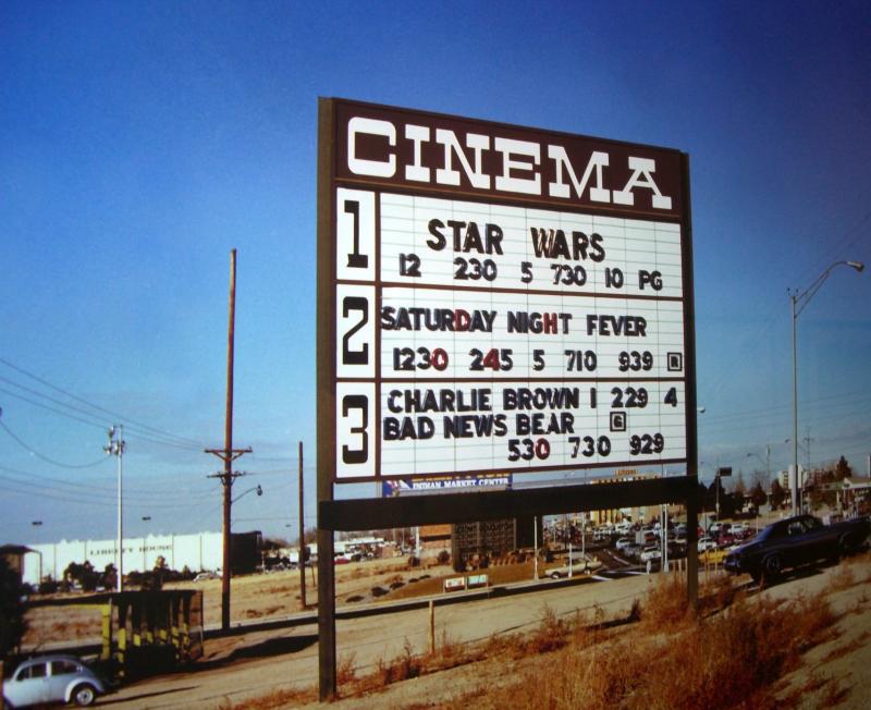 A cinema marquee sign displays showtimes for “Star Wars,” “Saturday Night Fever,” “Charlie Brown,” and “Bad News Bear,” set against a clear sky and roadside scene.
