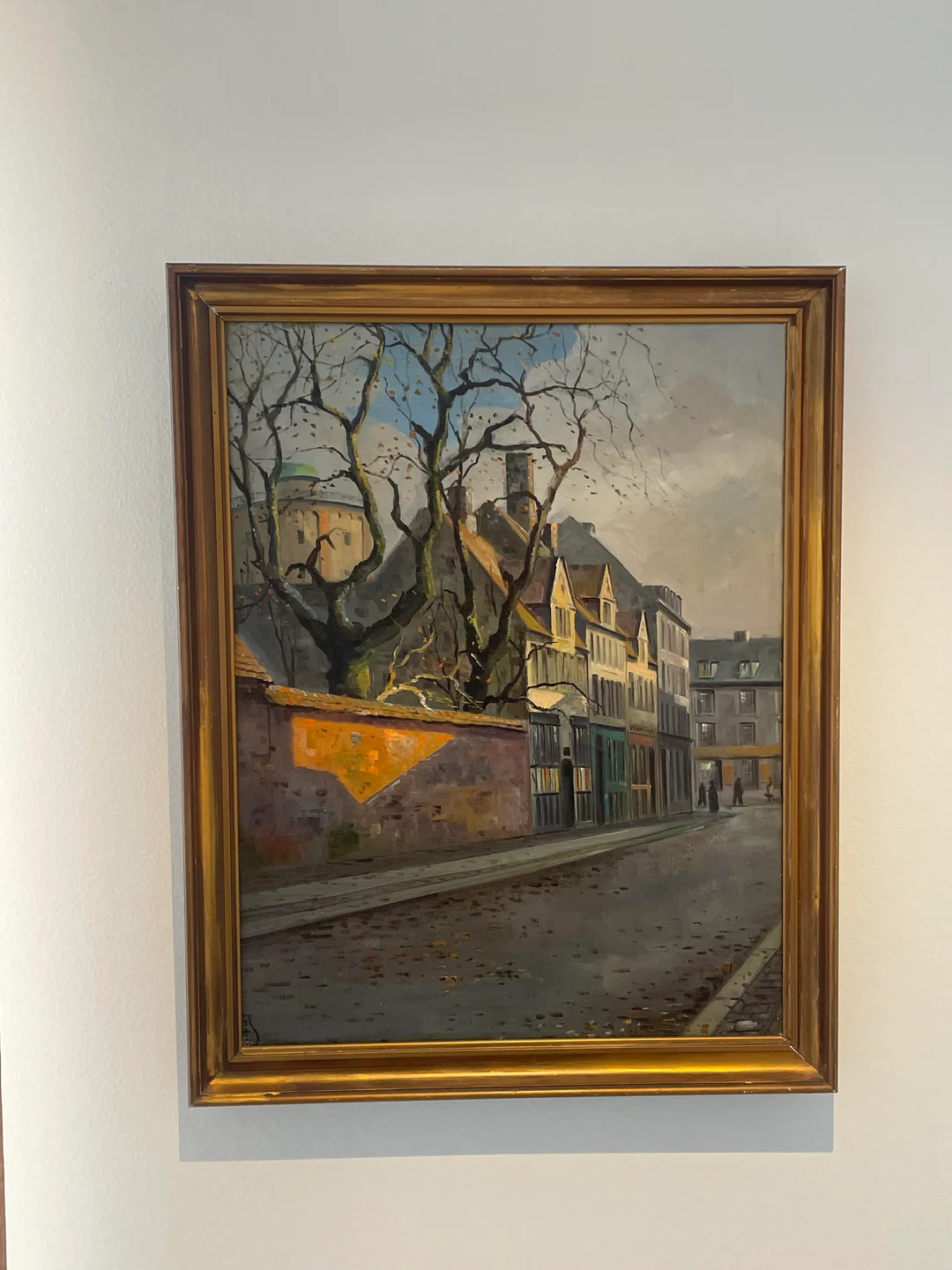 A framed painting of a quiet street with old buildings, leafless trees, a stone wall, and two small figures walking. The scene appears to be set in the late afternoon or early evening.