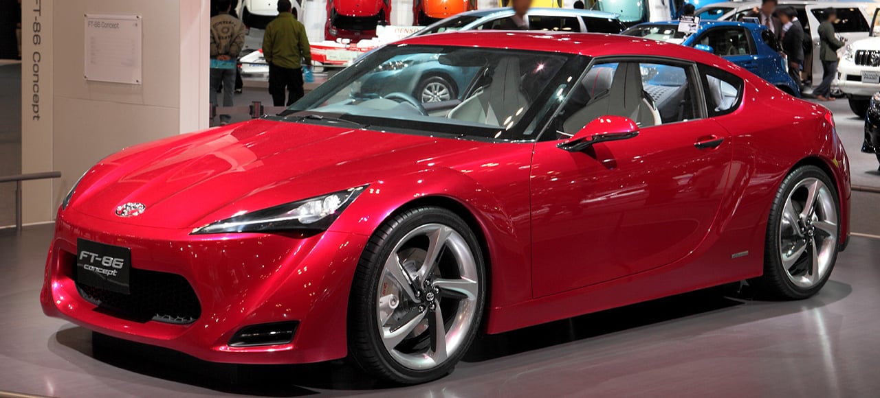 A sleek red sports car with a low profile and silver wheels is displayed indoors at an auto show, surrounded by other vehicles and people in the background.