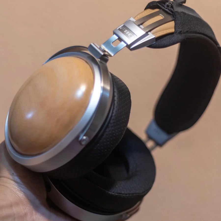 A close-up of a hand holding wooden-cup over-ear headphones with a black padded headband and metal accents, shown against a beige background.