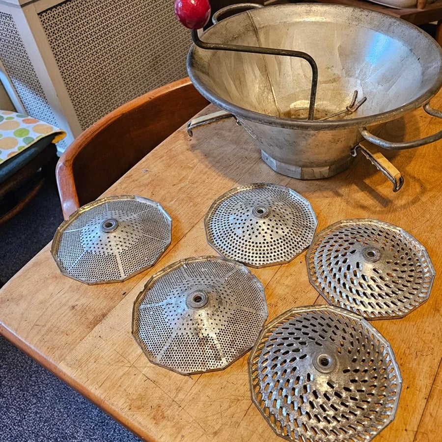 A vintage metal food mill with a red-handled crank is on a wooden table, along with five round interchangeable discs with different hole patterns.