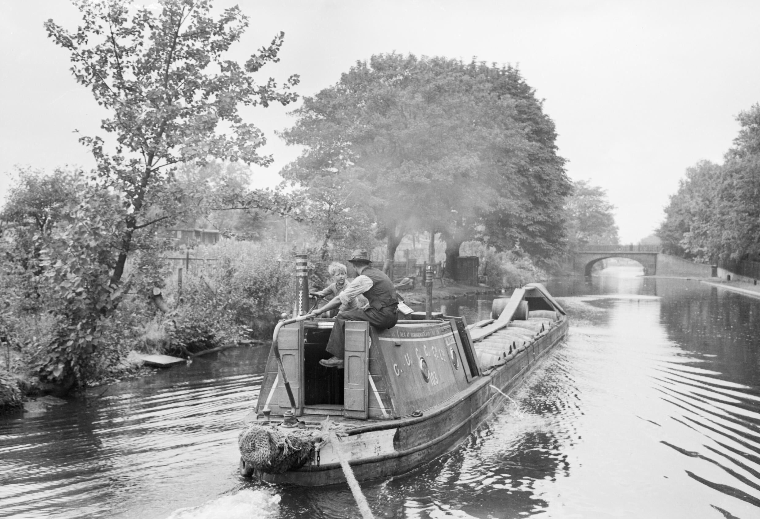 A man and a boy steer a narrowboat along a tree-lined canal. The boat creates ripples in the water, and a bridge is visible in the distance under a cloudy sky.