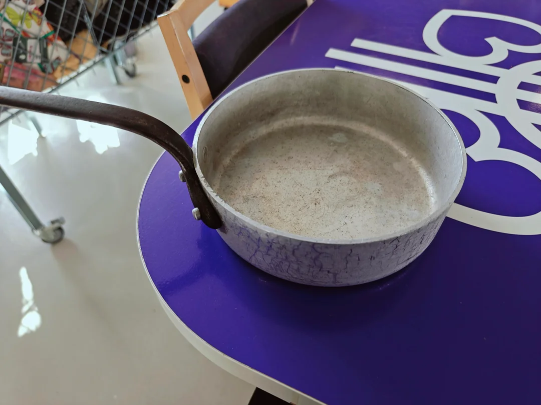 A worn, empty metal frying pan with a dark handle sits on a purple and white table. Part of a chair and a shopping cart are visible in the background.