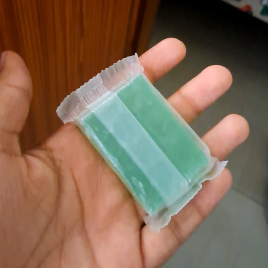 A hand holds a small, rectangular green soap bar wrapped in transparent plastic packaging, with a wooden surface and floor in the background.