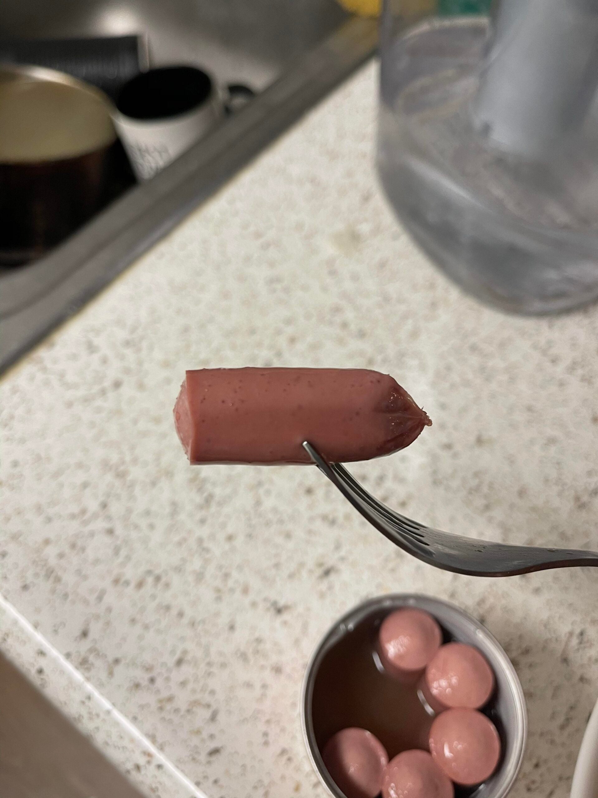 A fork holds a small sausage above a countertop, with an open can of mini sausages and a kitchen sink visible in the background.