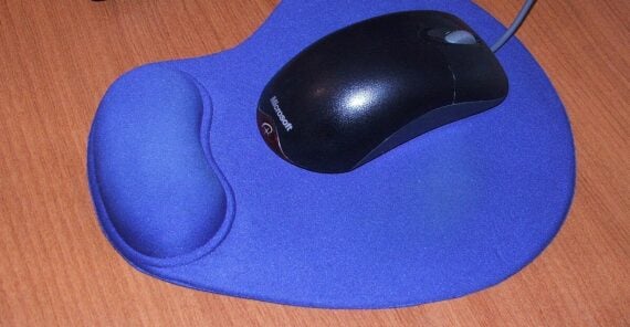 A black wired computer mouse on a blue mouse pad with a built-in wrist rest, placed on a wooden desk.