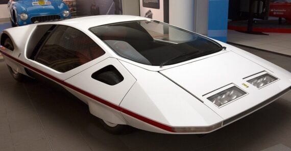 A futuristic, wedge-shaped white concept car with angular lines and large glass windows is displayed indoors on a tiled floor.