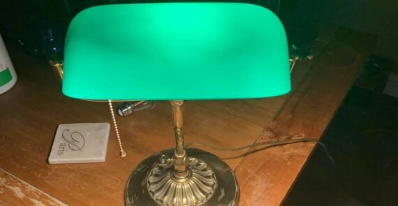 A green-shaded banker’s lamp with a brass base sits on a wooden desk, illuminated in a dark room.